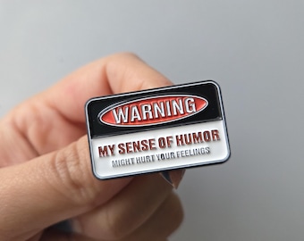 May include: A rectangular enamel pin with a black border and the word "WARNING" in red. The pin reads "MY SENSE OF HUMOR MIGHT HURT YOUR FEELINGS" in white text.