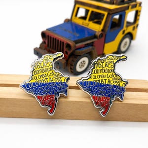 May include: Two pins in the shape of Colombia's map, with the country's flag colors and text. A wooden toy jeep in the background is painted in the colors of the Colombian flag. The pins are on a wooden stand.
