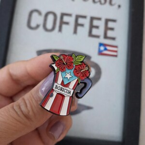 Pode incluir: Pin de esmalte com um design de cafeteira vermelha, branca e azul com flores de hibisco e a palavra "BORICUA". O pin tem um design da bandeira de Porto Rico e est&aacute; sobre um fundo desfocado com as palavras "But First, COFFEE".