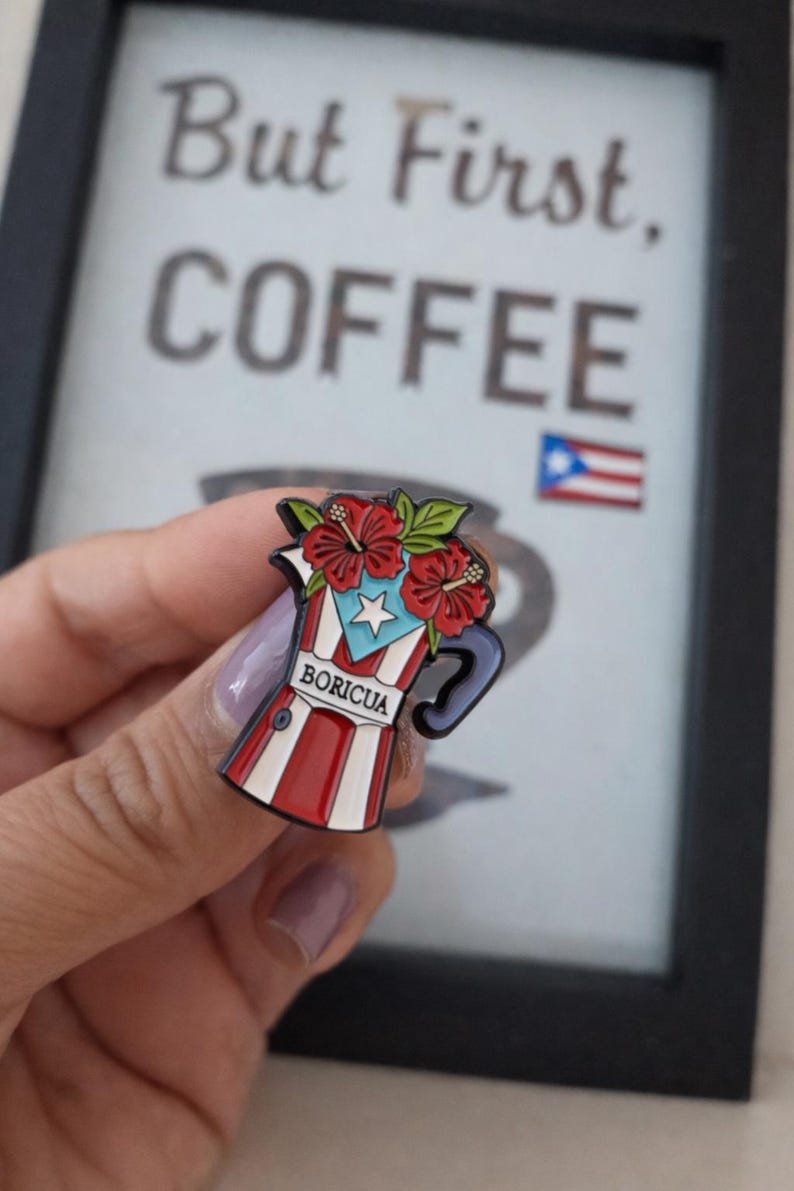 Pode incluir: Pin de esmalte com um design de cafeteira vermelha, branca e azul com flores de hibisco e a palavra "BORICUA". O pin tem um design da bandeira de Porto Rico e est&aacute; sobre um fundo desfocado com as palavras "But First, COFFEE".