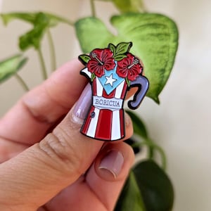 Pode incluir: Um alfinete de esmalte colorido em forma de cafeteira, com a bandeira de Porto Rico e flores de hibisco vermelhas. O alfinete tem um esquema de cores vermelho, branco e azul com a palavra "BORICUA" exibida de forma proeminente.