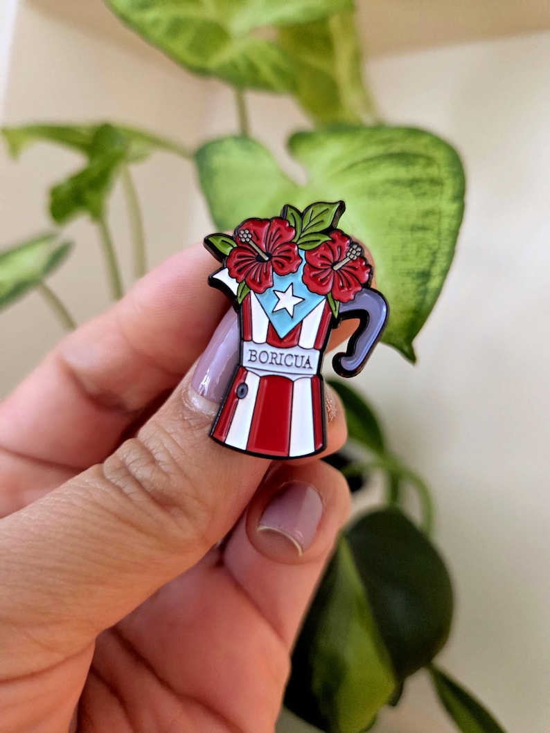 Pode incluir: Um alfinete de esmalte colorido em forma de cafeteira, com a bandeira de Porto Rico e flores de hibisco vermelhas. O alfinete tem um esquema de cores vermelho, branco e azul com a palavra "BORICUA" exibida de forma proeminente.