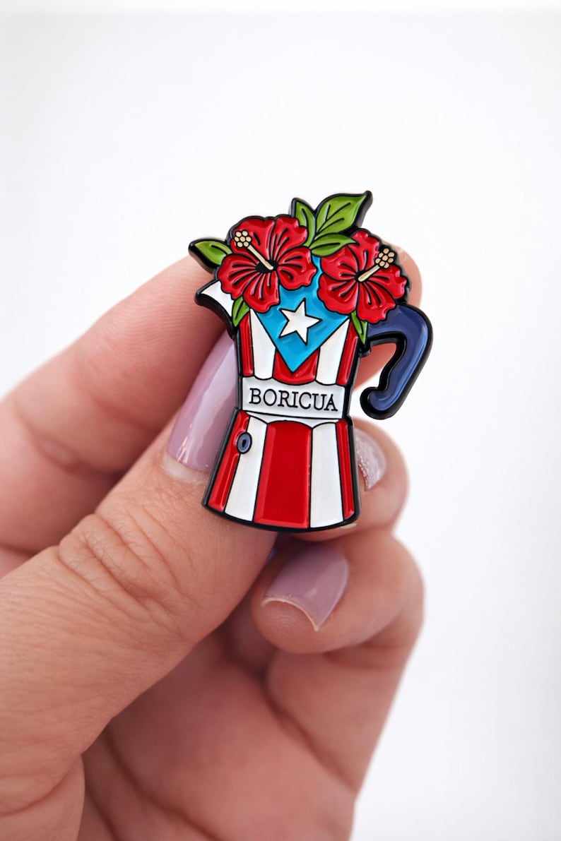 May include: A colorful enamel pin in the shape of a coffee pot, featuring the Puerto Rican flag design. The pin is adorned with red hibiscus flowers and green leaves. The word "BORICUA" is printed on the pin.