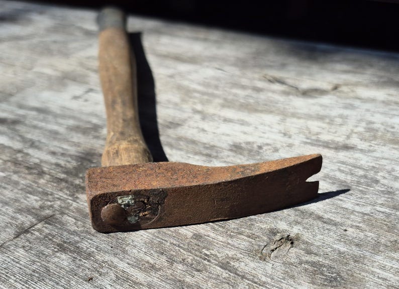 May include: A rusty, vintage claw hammer with a wooden handle. The hammer head is a dark, weathered brown, showing signs of age and use. The wooden handle is a light brown color. The tool is resting on a weathered wooden surface.
