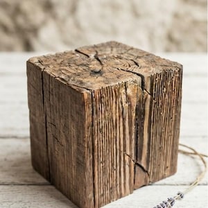 May include: A square block of weathered brown wood with a rough, textured grain and visible cracks on the top surface. A thin piece of twine with a small sprig of dried purple lavender is attached to the side of the wooden block.
