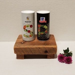 May include: Two spice containers, one white with red and green illustrations and the text "MORTON" and the other black with green and white illustrations and the text "McCormick", are sitting on a wooden stand.