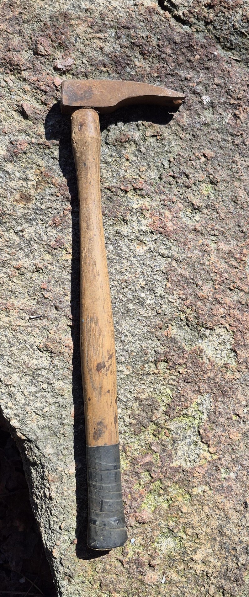 May include: A well-worn claw hammer with a wooden handle and a metal head. The hammer is resting on a rough, textured rock surface. The handle shows signs of use.