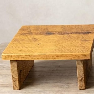May include: A square wooden riser with a natural light brown finish and visible wood grain patterns. The stand features a flat top surface supported by two vertical side panels, creating a low-profile platform for display purposes.