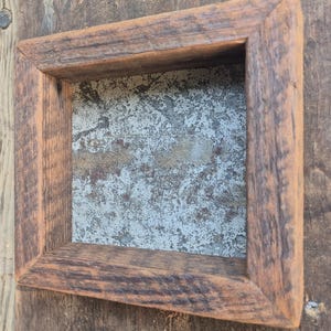 May include: A square frame constructed from weathered brown wood with a visible grain texture. The center of the frame features a flat, rectangular panel made of gray metal with a mottled, distressed surface texture showing patches of oxidation.