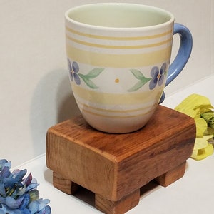 May include: A yellow and white striped mug with blue flowers and a blue handle sits on a wooden coaster with four legs.