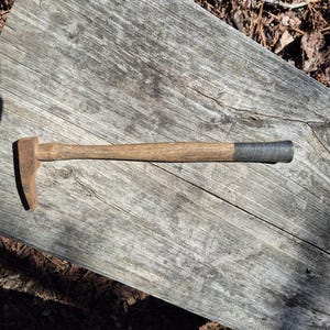 May include: A vintage hammer with a wooden handle and a metal head. The handle is light brown, and the head is a rusty brown color. The end of the handle is wrapped in a dark gray material. The hammer is lying on a weathered wooden surface.