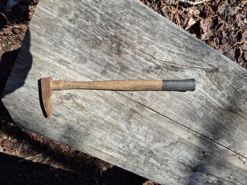 May include: A vintage hammer with a wooden handle and a metal head. The handle is light brown, and the head is a rusty brown color. The end of the handle is wrapped in a dark gray material. The hammer is lying on a weathered wooden surface.