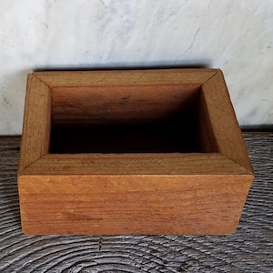 May include: A rectangular wooden box with a natural brown finish. The box has a simple design with a smooth surface and a slightly raised edge around the opening.