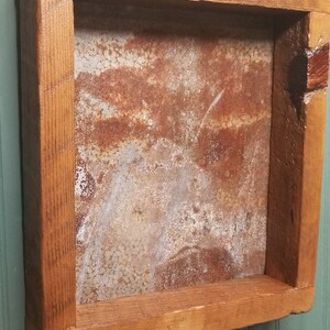 May include: A rustic wooden frame with a square opening, showcasing a piece of rusty metal with a mottled, aged texture. The frame is a rich brown color with visible wood grain.