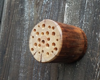May include: A wooden bee house with a circular opening and multiple smaller holes drilled into the wood. The bee house is attached to a wooden wall.