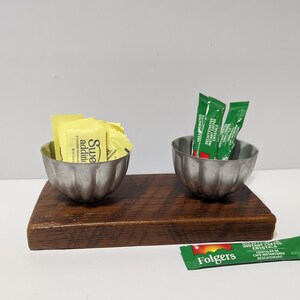 May include: Two small silver bowls with a wooden base. One bowl contains yellow sugar packets, the other contains green coffee creamer packets. The text on the packets reads "Sweet'n Low", "Decaffeinated Instant Coffee Crystals", and "Folgers Decaffeinated Instant Coffee Crystals".