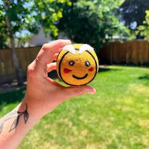 May include: A round, yellow needle-felted bee toy featuring black stripes, white felt wings on top, and a stitched face with black eyes, a curved black mouth, and small red cheeks. The textured wool surface shows detailed craftsmanship.