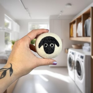 May include: A white wool dryer ball with a black sheep face design. The ball is being held in a hand.
