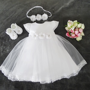 Pu&ograve; includere: Abito bianco per neonata con gonna in tulle e bordino in pizzo. L'abito ha un corpetto in raso con un ricamo floreale. Il set include una fascia per capelli e scarpe abbinate.