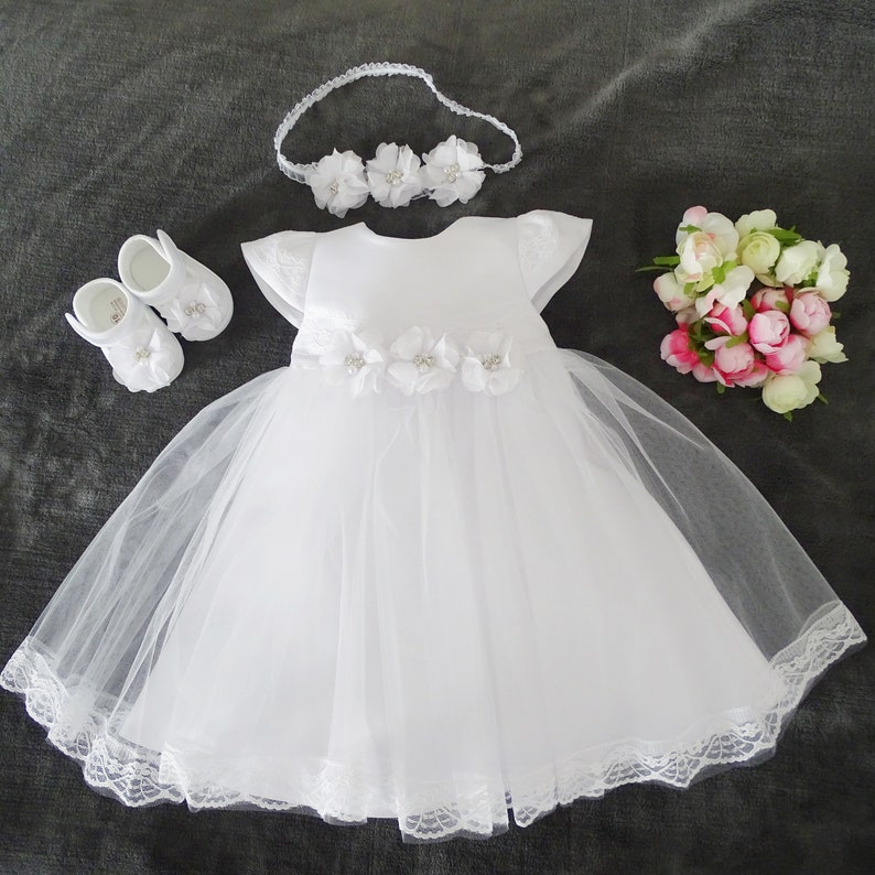 Pu&ograve; includere: Abito bianco per neonata con gonna in tulle e bordino in pizzo. L'abito ha un corpetto in raso con decorazioni di fiori bianchi. Il set include una fascia per capelli e scarpe abbinate.