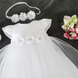 Pu&ograve; includere: Abito bianco per neonata con gonna in tulle e corpetto in raso. L'abito &egrave; decorato con un bordo in pizzo e tre fiori bianchi con strass sul corpetto. &Egrave; incluso un cerchietto bianco con tre fiori bianchi e strass.