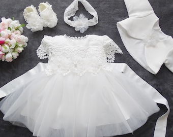 Può includere: Abito bianco per neonata con dettagli in pizzo, una fascia per capelli abbinata con un fiore e una giacca bolero bianca. L'abito ha una gonna in tulle e un nastro di raso.