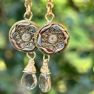 May include: Gold earrings with a round, ornate, multi-colored design and a clear teardrop-shaped crystal pendant.