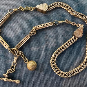 May include: An antique silver watch chain with a decorative T-bar, a textured ball, and a series of rectangular links with cut-out designs. The chain also features a thick curb chain section and decorative clasps.