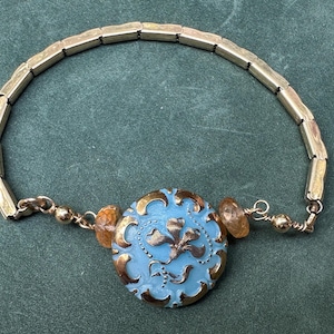 May include: A bracelet featuring a central round light blue glass button with gold-toned floral detailing. The button is flanked by two small amber-colored faceted beads and a gold-toned metal chain composed of rectangular links and small spheres.