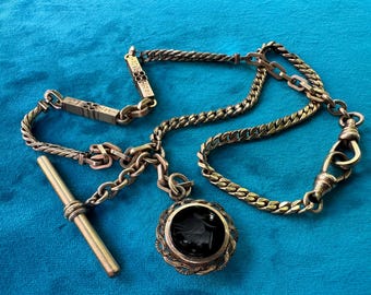 Puede incluir: Cadena de reloj de bolsillo dorada antigua con un adorno decorativo y un colgante redondo con un camafeo negro. La cadena tiene una variedad de estilos de eslabones, incluyendo una cadena de bordillo y una cadena de cuerda trenzada.