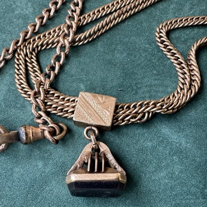 May include: A gold chain with a decorative pendant. The pendant has a square top with an engraved design and a black stone at the bottom.