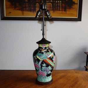 May include: A black ceramic table lamp with a colorful bird and floral design. The lamp has a dark brown base and a black lampshade. The lamp is on a wooden table.