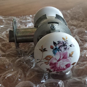 May include: A white porcelain doorknob with a floral design in pink, purple, blue, and gold. The doorknob is part of a silver-colored door lock mechanism. The knob is resting on bubble wrap.