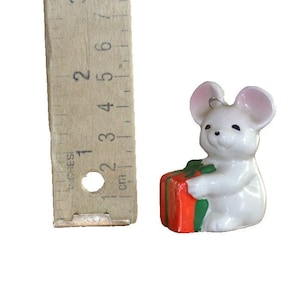 May include: A small, white ceramic mouse figurine with pink-lined ears, holding a red and green gift box. The figurine is approximately 1.5 inches tall. A wooden ruler is next to the mouse.