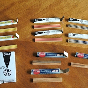 May include: An assortment of vintage Scripto pencil lead boxes and individual leads. The boxes are white with black and red accents, and the text "Scripto Smooth Writing LEADS" is visible. The leads are in various colors.