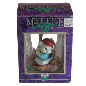 May include: A white bear figurine wearing a red hat and blue sweater inside a purple MISTLETOE MAGIC COLLECTION box. The box features a clear plastic window, a gold hanging string, and a decorative mistletoe graphic at the bottom center.