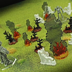 May include: Miniature tabletop game terrain set featuring a gray robot figure, white building ruins, black trees, and translucent orange flames and smoke. The scene is set on a green surface.