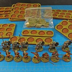 May include: A collection of miniature figurines and bases for tabletop gaming. The figurines are painted in various colors, including brown, gold, and red. The bases are brown with circular indentations. A bag of green tokens is also included.
