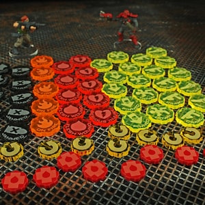 May include: A set of colorful plastic tokens for a tabletop game. The tokens are in various shapes and sizes, and feature different symbols and text, including "Out of Ammo", "Jammed", "On Fire", "Flesh Wounds", "Critical", "Pinned", "Down", and "Crank".