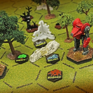 May include: A tabletop wargaming scene featuring a red mech and a blue mech on a green hexagonal grid map. The board includes miniature trees, a white ruin piece, a gray rock, and black hexagonal tokens labeled Rubble, Heavy, Rough, Light, and Bogged.