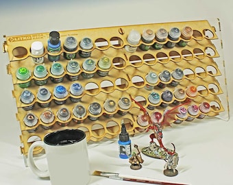Pode incluir: Um suporte de madeira para frascos de tinta com vários frascos de tinta de várias cores. Uma caneca branca com interior preto, um pincel e miniaturas estão em frente ao suporte. O suporte tem o texto "LITKO MEGA RACK".