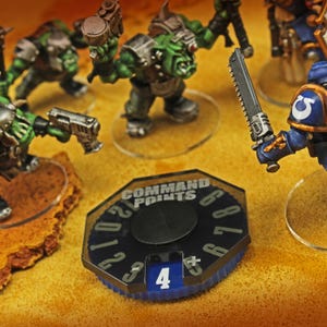 May include: A tabletop game command points counter with the text "COMMAND POINTS" and numbers 0-9. The counter is black with a blue rim and a dial showing the number 4. Miniature figures of green-skinned orcs and blue-armored soldiers are in the background.