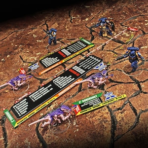 May include: Miniature figurines of blue-armored soldiers and purple creatures are positioned on a textured, brown surface. Game cards with text like "ATTACKS" and "SHOOTING PHASE" are scattered around the figures. The scene suggests a tabletop war game.