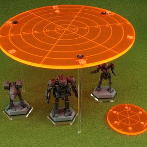 May include: Orange plastic circular template with a grid pattern and concentric circles. Three miniature robots are standing on hexagonal bases. The template is suspended above the robots on clear acrylic rods.