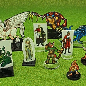 May include: A collection of fantasy character figurines, including a winged horse, a green monster, and various humanoids. The figures are made of cardboard and plastic, with detailed illustrations and stand-up bases. The background is a green surface.