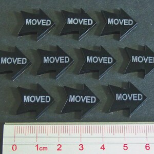 May include: Ten black arrow-shaped stickers with the word "MOVED" in white. A ruler is at the bottom, showing measurements in centimeters. The stickers are arranged in three rows.