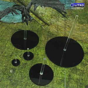May include: A collection of black, circular display bases with clear acrylic stems, designed for tabletop gaming miniatures. The bases vary in size, with a dragon miniature mounted on one. The background is a green and brown game mat.