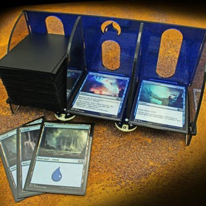 May include: A blue and black card holder with three compartments, holding a stack of black cards and several Magic: The Gathering cards. The holder has a curved design with cut-out shapes and is set on a textured surface.