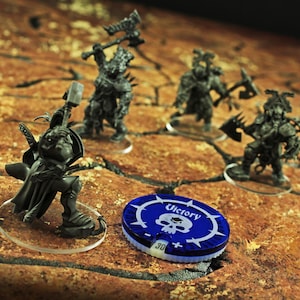 May include: Miniature figurines of fantasy warriors with weapons, posed on clear bases, are arranged on a textured brown surface. A blue and white game token with a skull and the word "Victory" is also present. The figures are dark gray.