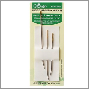 May include: A package of Clover Huck Embroidery Needles, Art No. 2012. The package is yellow with green accents and features the Clover logo. The needles are displayed on a white background. The package includes instructions in English, French, Spanish, German, and Dutch.